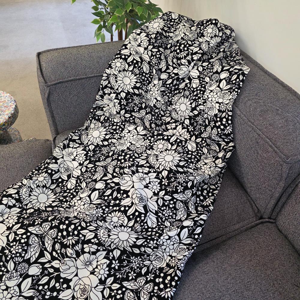 Black and white uncolored vintage floral and butterfly patterned blanket with a black fleece lined underside draped over a gray couch.