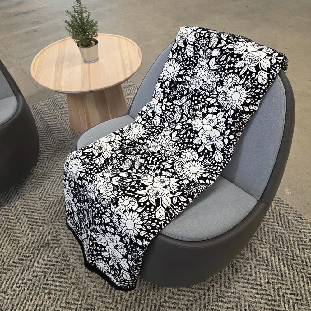 Black and white uncolored vintage floral and butterfly patterned blanket with a black fleece lined underside draped over a gray chair.