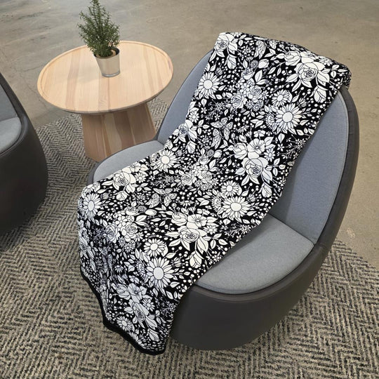 Black and white uncolored vintage floral and butterfly patterned blanket with a black fleece lined underside draped over a gray chair.