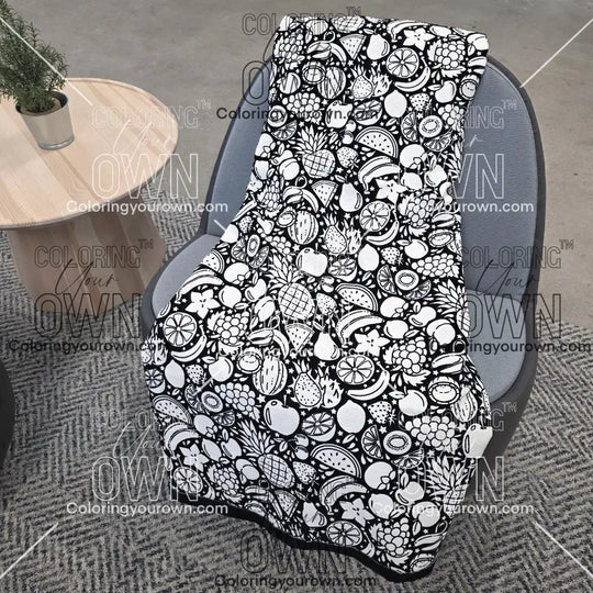 Black and white uncolored fruit variety with pineapples, bananas, watermelons and more patterned blanket with a black fleece lined underside draped over a gray chair.