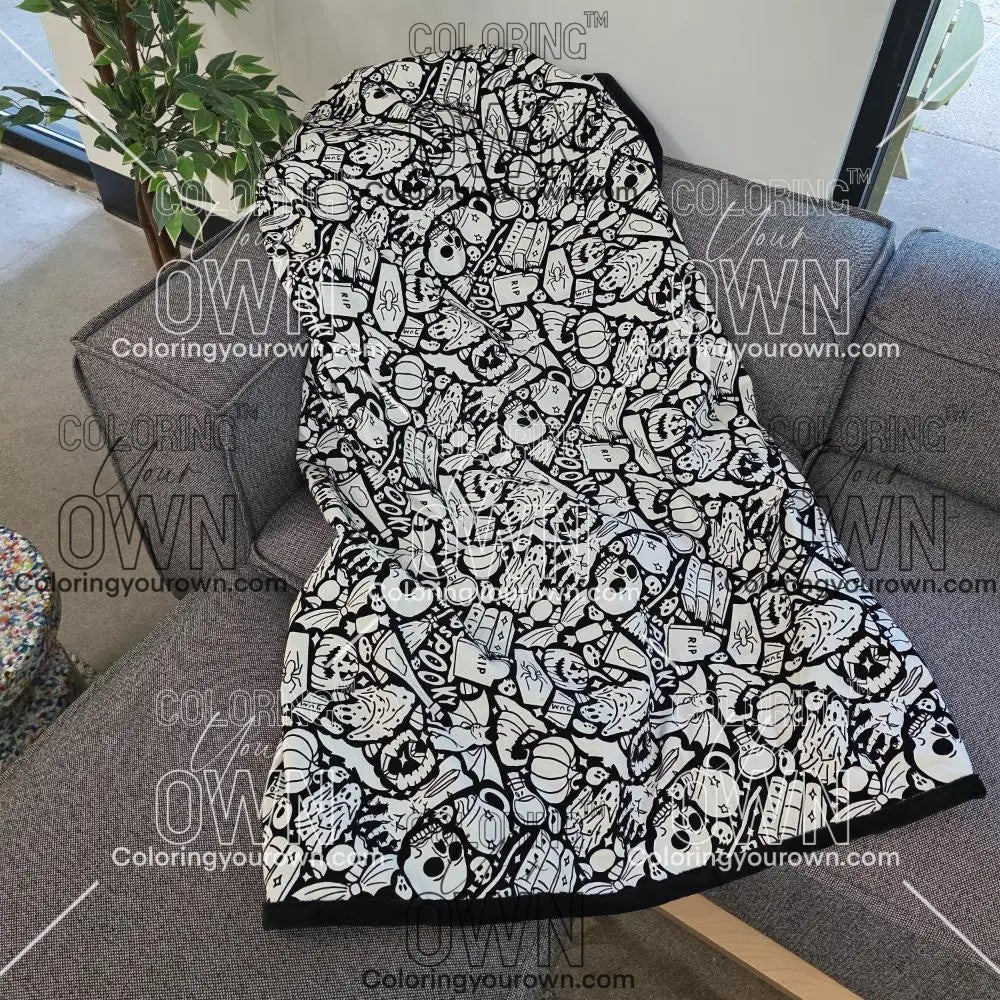 Black and white uncolored spooky halloween patterned blanket with skulls and bats and a black fleece lined underside draped over a gray couch.