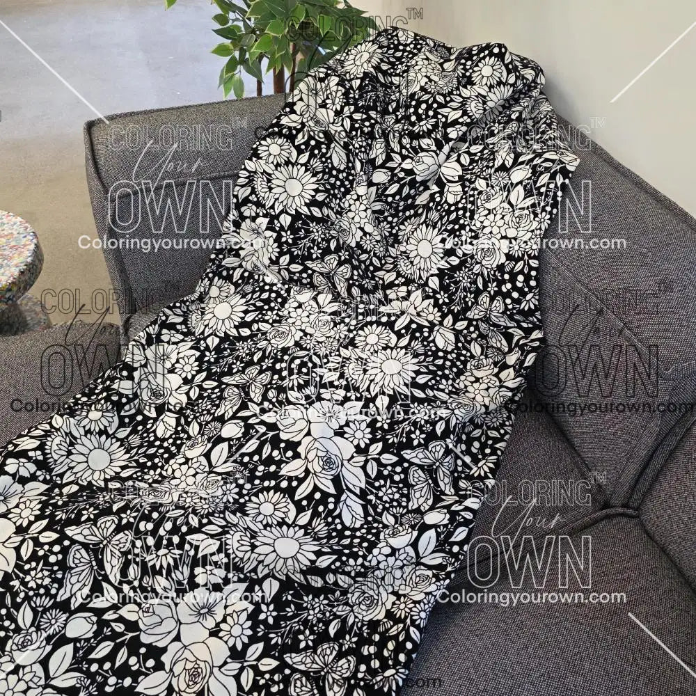 Black and white uncolored vintage floral and butterfly patterned blanket with a black fleece lined underside draped over a gray couch.