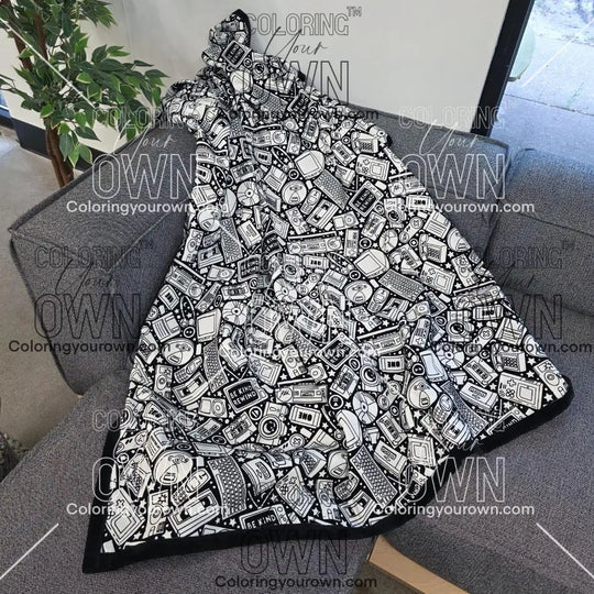 Black and white uncolored retro technology, keyboard ,camera and stereo patterned blanket with a black fleece lined underside draped over a gray couch.