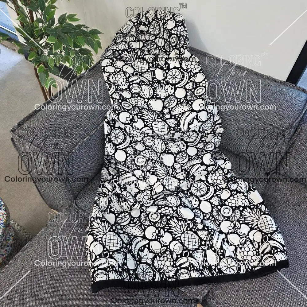 Black and white uncolored fruit variety with pineapples, bananas, watermelons and more patterned blanket with a black fleece lined underside draped over a gray couch.