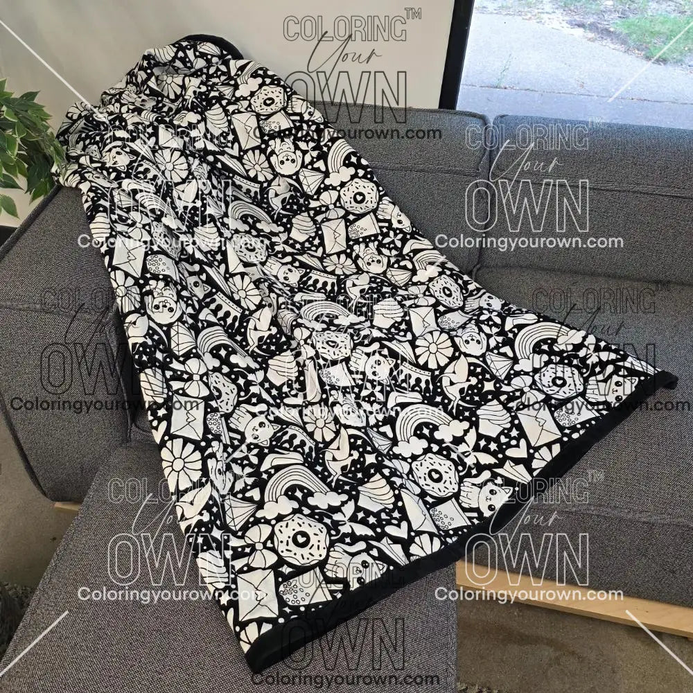 Black and white uncolored version two doodle patterned blanket with donuts, cats, cupcakes a black fleece lined underside draped over a gray couch.