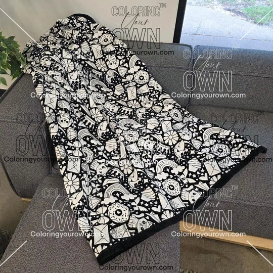 Black and white uncolored version two doodle patterned blanket with donuts, cats, cupcakes a black fleece lined underside draped over a gray couch.