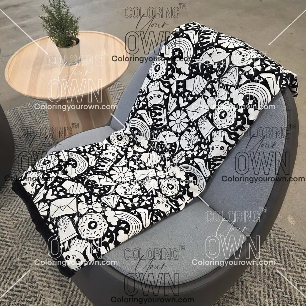 Black and white uncolored version two doodle patterned blanket with donuts, cats, cupcakes  a black fleece lined underside draped over a gray chair.