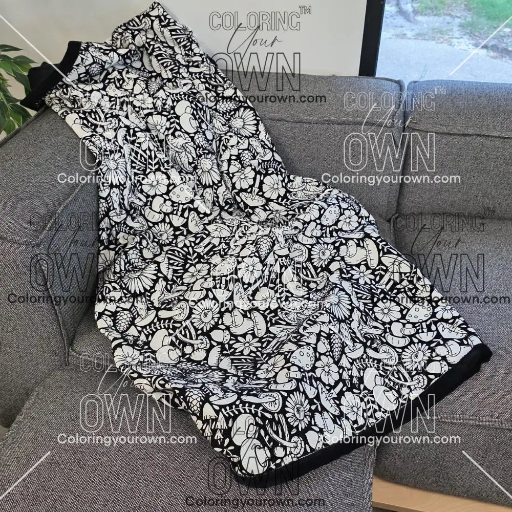 Black and white uncolored mushroom and floral patterned blanket with black fleece lined underside draped over a gray couch.