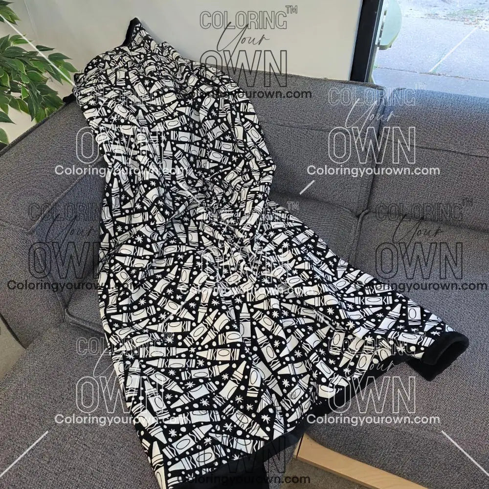 Black and white uncolored many crayon patterned blanket with a black fleece lined underside draped over a gray couch.