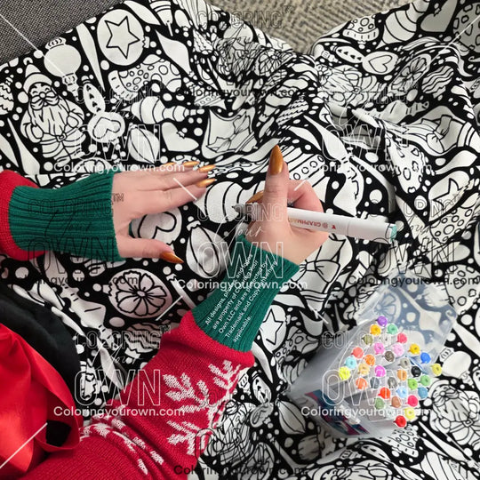 Different angle of a close-up of a black-and-white coloring blanket with a Christmas ornament-themed line-art pattern as a person colors the design with it draped over her lap. Coloring Your Own branding is all over the photo in the form of a watermark.