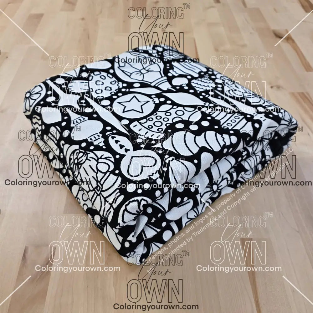 Folded black-and-white Coloring Your Own blanket featuring a Christmas ornament–themed line-art pattern, displayed on a light wood surface.