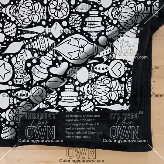 Close-up of a black-and-white coloring blanket showing a Christmas-themed ornament line-art pattern. One corner of the blanket is folded over, partially obscuring the design. “Coloring Your Own” branding is visible along the edge.
