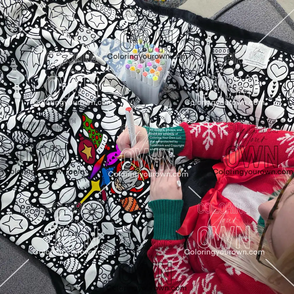 Close-up of a black-and-white coloring blanket with a Christmas ornament-themed line-art pattern as a person colors the design with it draped over her lap. Coloring Your Own branding is all over the photo in the form of a watermark.