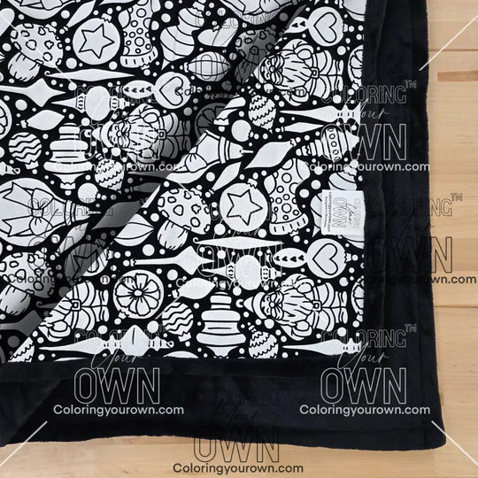 Close-up of a black-and-white coloring blanket showing a Christmas Ornament-themed line-art pattern. One corner of the blanket is folded over, partially obscuring the design. This fold is slightly higher than the fold in another photo displayed in this  product listing.  “Coloring Your Own” branding is visible along the edge on the tag of the blanket. "Coloring Your Own" branding is also over the whole photo as a watermark.