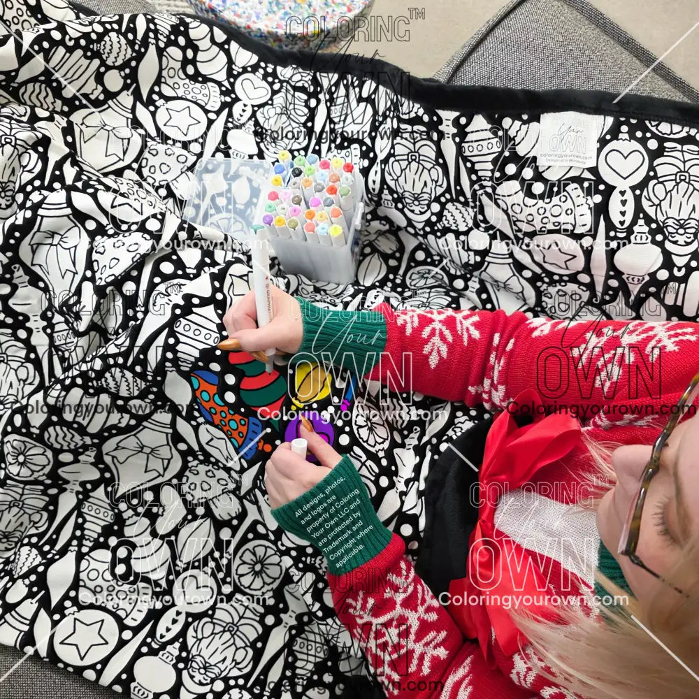 A third angle of a close-up of a black-and-white coloring blanket with a Christmas ornament-themed line-art pattern as a person colors the design with it draped over her lap. Coloring Your Own branding is all over the photo in the form of a watermark.