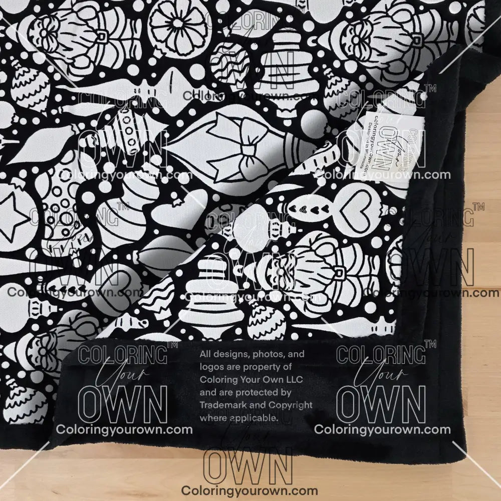 Close-up of a black-and-white coloring blanket showing a Christmas-themed ornament line-art pattern. One corner of the blanket is folded over, partially obscuring the design. “Coloring Your Own” branding is visible along the edge.