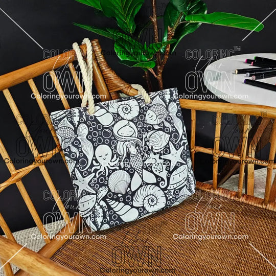photo of an uncolored color your own tote sitting on a wicker chair beside a table with markers
