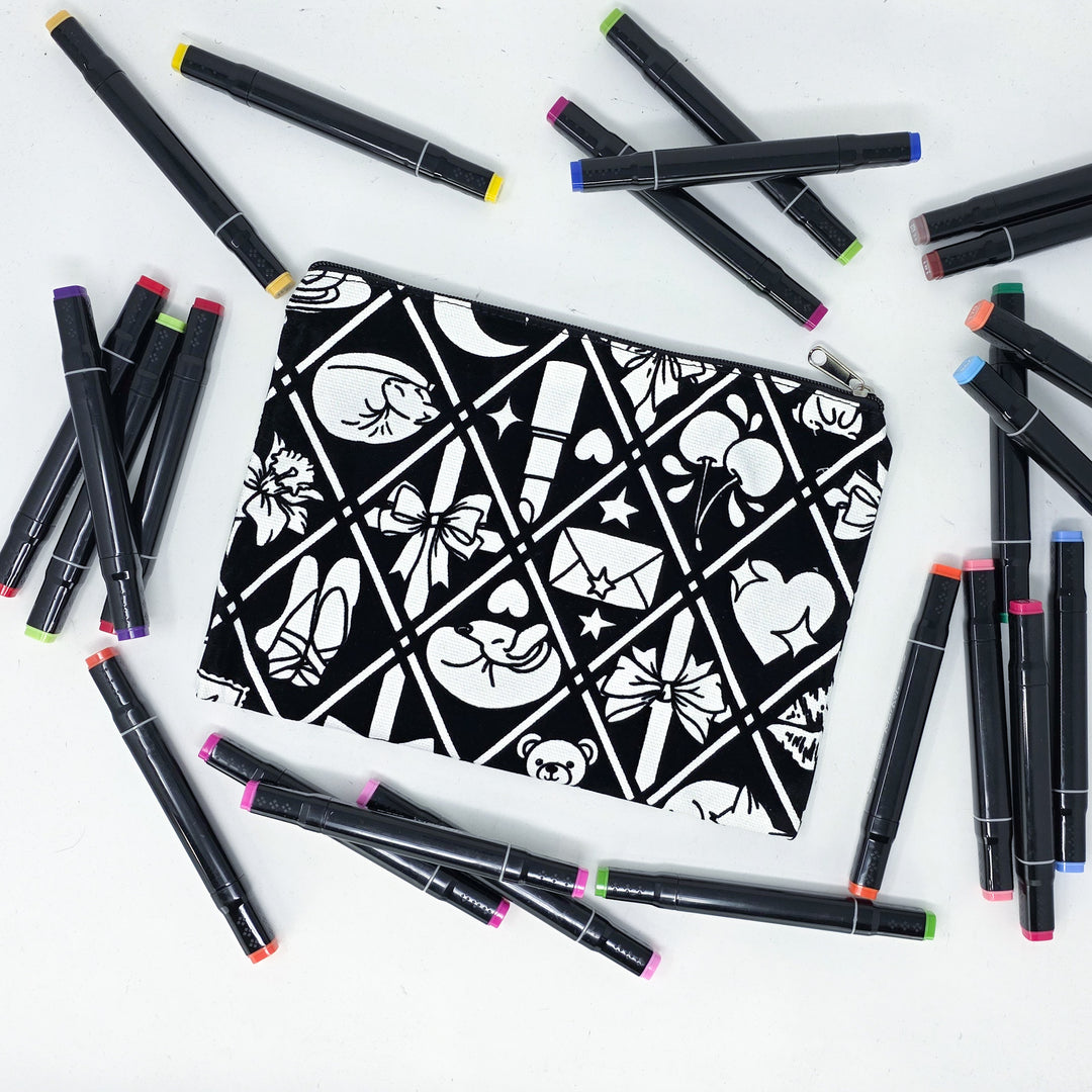 Photo of a black and white flat pencil pouch with lots of colorful markers beside it indicating the customer gets to color it when they receive theirs. This is the same bag as the pencil case and makeup bag - Side A - this bag has images of Coquette-inspired illustrations including a swan, a paw print, bows, a moon, stars, a bunny on it cherries and more