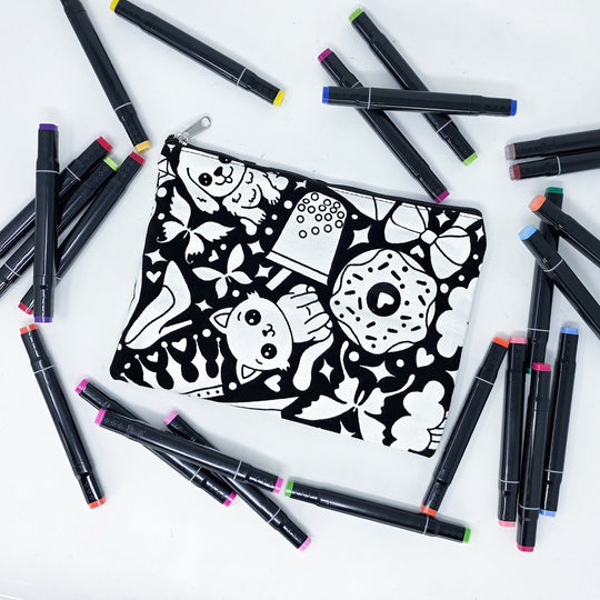 Photo of a black and white flat pencil pouch with lots of colorful markers beside it indicating the customer gets to color it when they receive theirs. This is the same bag as the pencil case - Side A - this bag has images of happy doodles on it