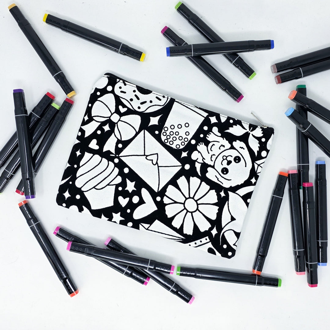 Photo of a black and white flat pencil pouch with lots of colorful markers beside it indicating the customer gets to color it when they receive theirs. This is the same bag as the pencil case and makeup bag - Side B - this bag has images of happy doodles on it