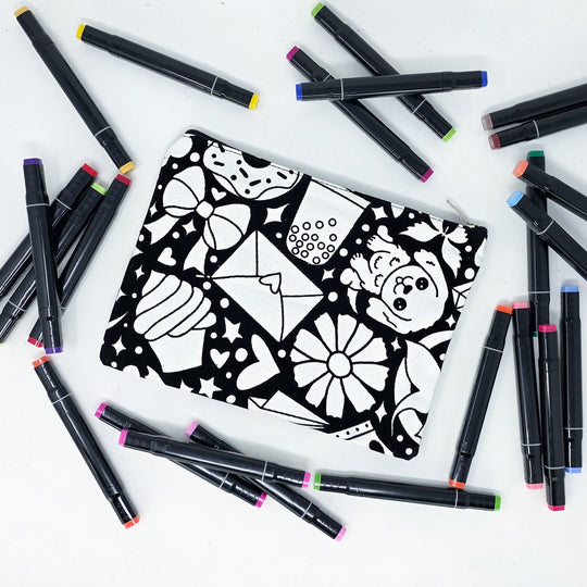 Photo of a black and white flat pencil pouch with lots of colorful markers beside it indicating the customer gets to color it when they receive theirs. This is the same bag as the pencil case and makeup bag - Side B - this bag has images of happy doodles on it