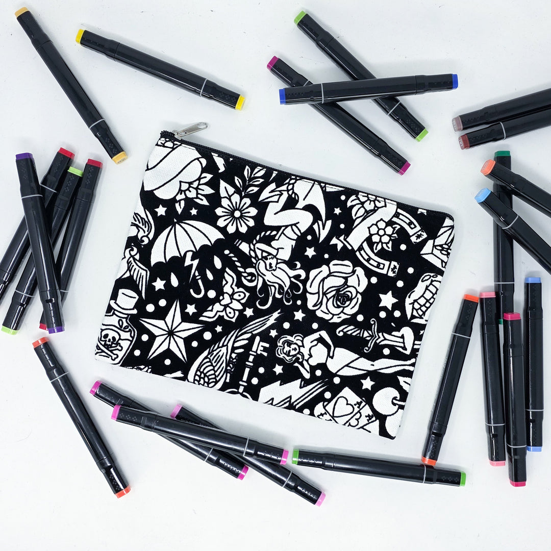 Photo of a black and white flat pencil pouch with lots of colorful markers beside it indicating the customer gets to color it when they receive theirs. This is the same bag as the pencil case and makeup bag - Side B - this bag has images of traditional tattoo-inspired graphics including pinup girls, cigarettes, roses, birds, vampire lips, cards, poison bottles, and more