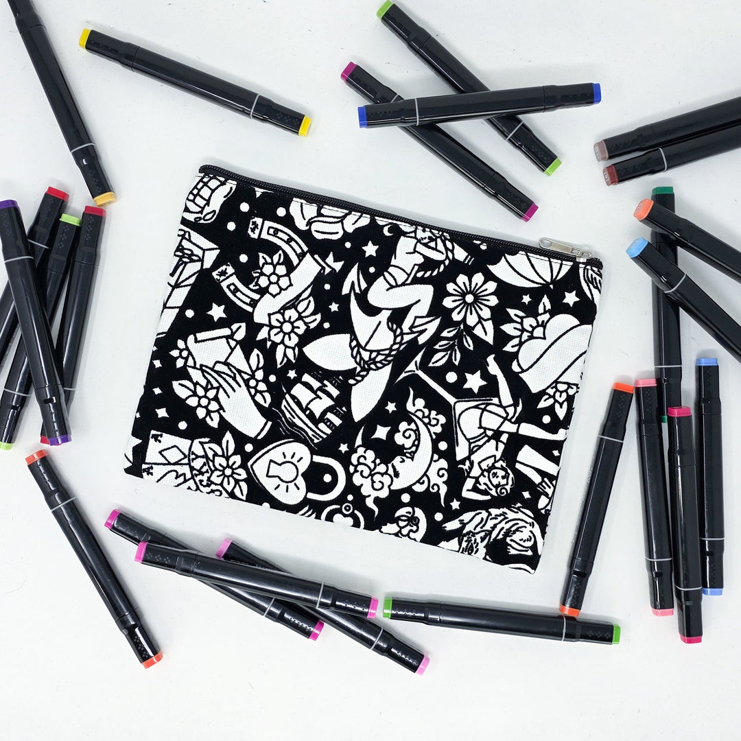 Photo of a black and white flat pencil pouch with lots of colorful markers beside it indicating the customer gets to color it when they receive theirs. This is the same bag as the pencil case and makeup bag - Side A - this bag has images of traditional tattoo-inspired graphics including pinup girls, cigarettes, roses, birds, vampire lips, cards, poison bottles, and more