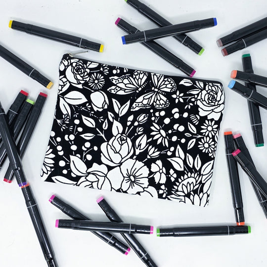 Photo of a black and white flat pencil pouch with lots of colorful markers beside it indicating the customer gets to color it when they receive theirs. This is the same bag as the pencil case - Side A - this bag has images of vintage flowers on it